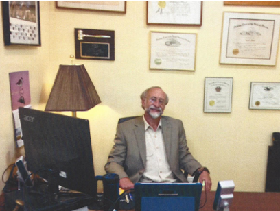 David Beale sits at his desk with his diplomas and framed certifications on the wall behind him
